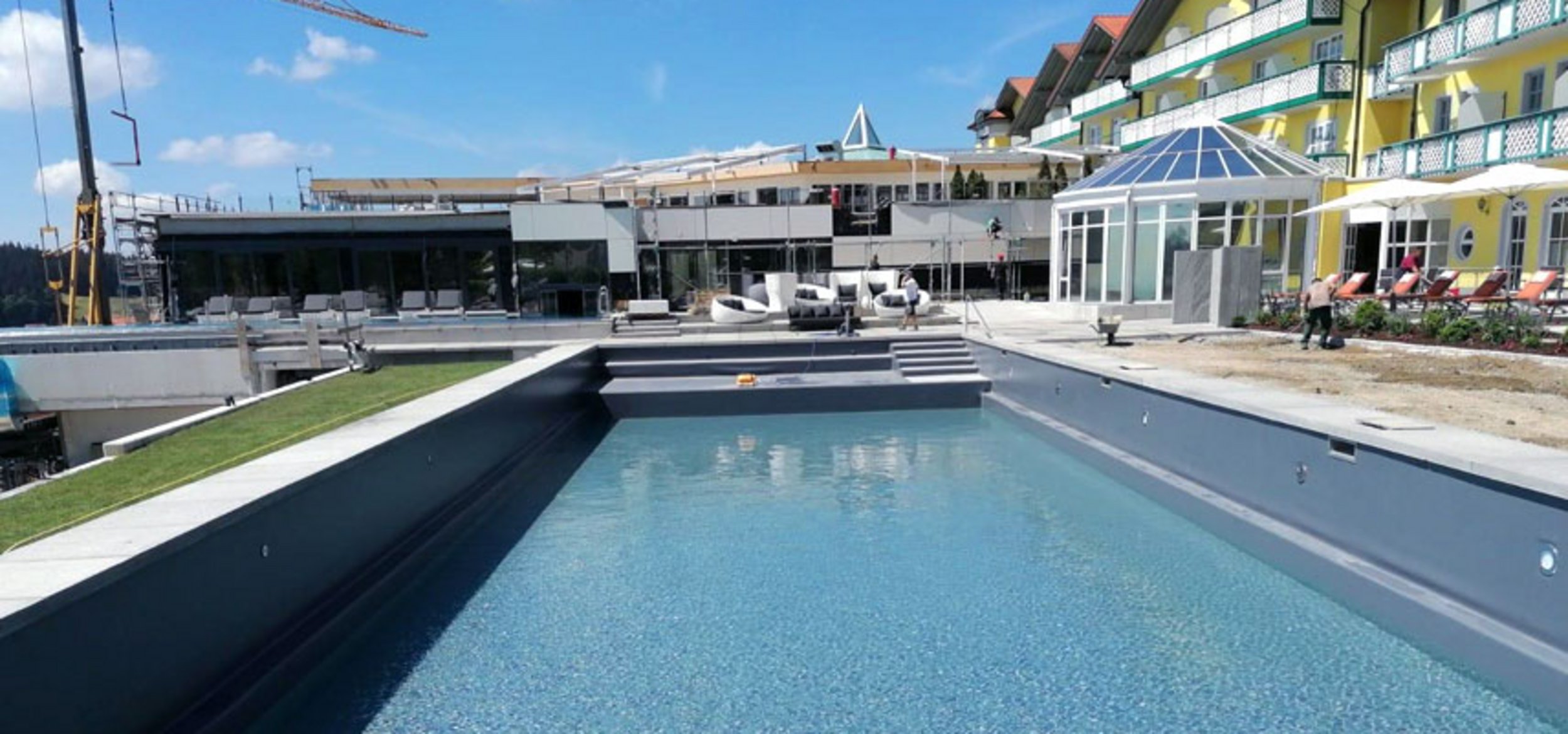 Blick vom Naturpool über die Pooltreppe auf das Hautpgebäude des Naturpool mit Liegen und Gästen im Sport- und Wellness­hotel „Angerhof” in St. Englmar
